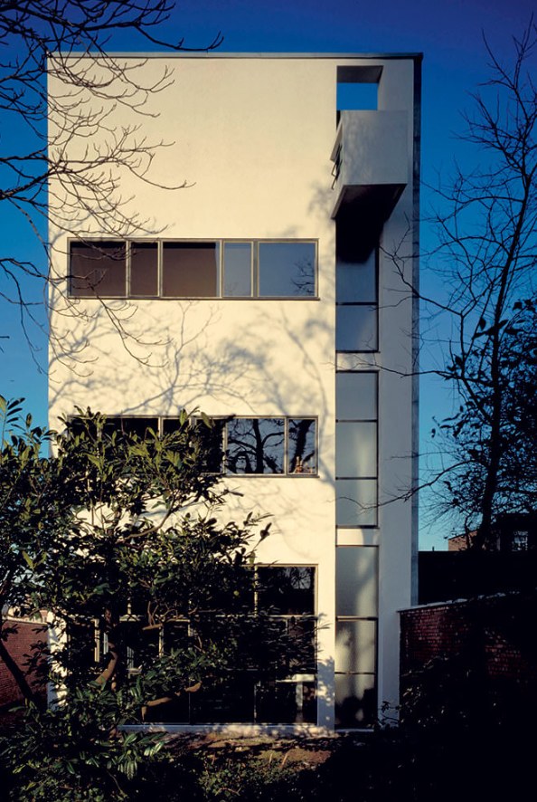 La fachada de la Casa Guiette a la calle, blanca y estrecha, contra
                un cielo azul de invierno: tres ventanas corridas, la franja
                vertical de la derecha y el vuelo del último piso. Delante, las
                ramas de un árbol sin hojas y un seto.